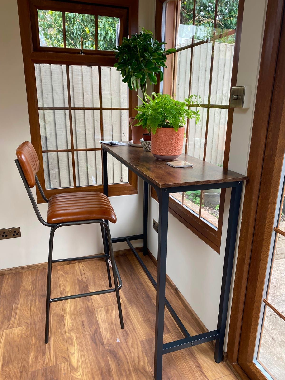 Garden room home office corner with a desk, leather stool and house plants beside hardwood windows
