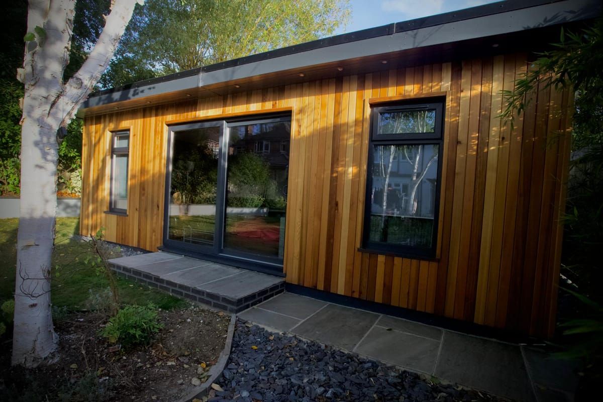 Cedar-clad garden room in daylight with anthracite sliding doors, beneath a silver birch