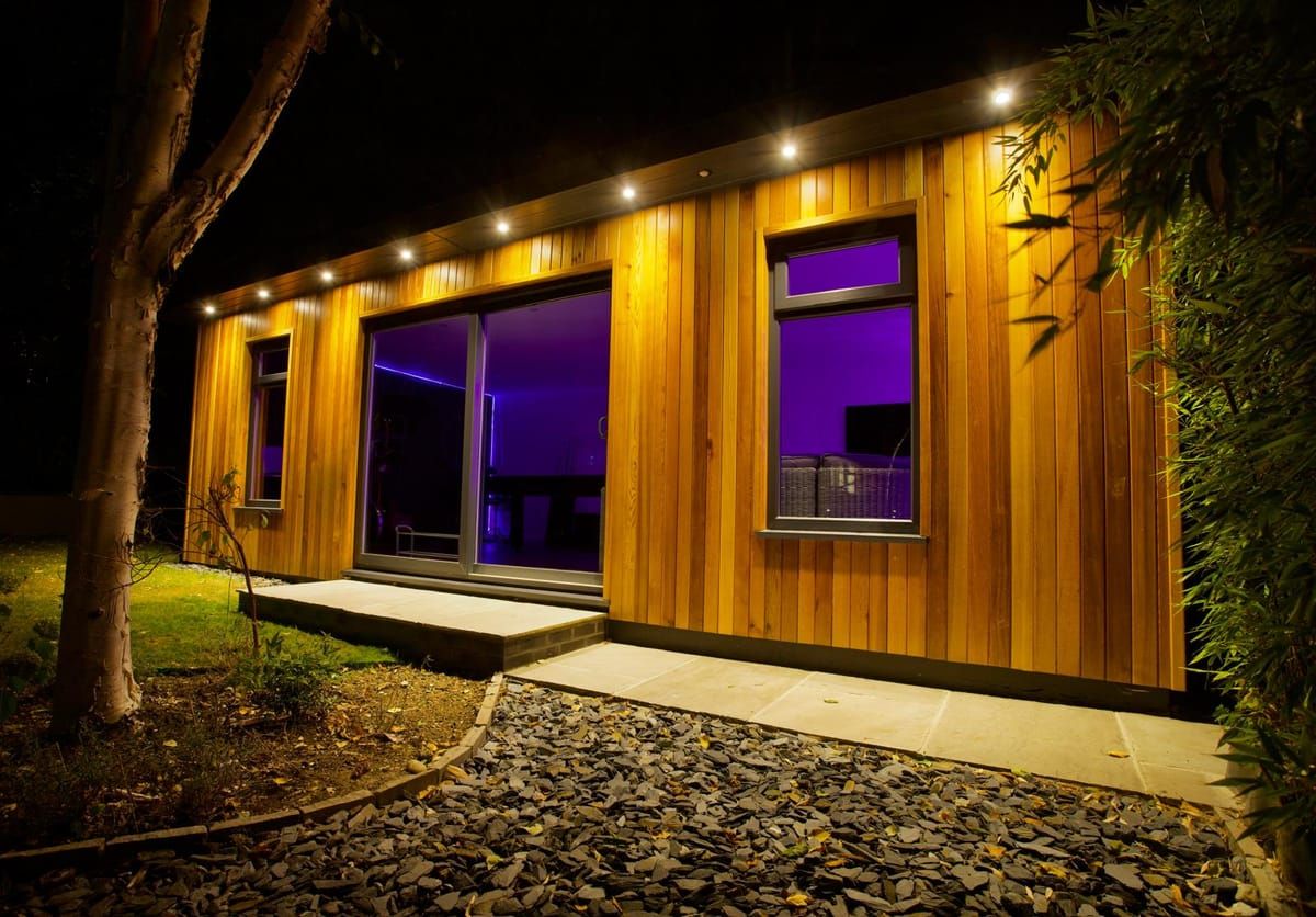 Cedar-clad garden games room lit at night, with recessed soffit downlights and a sandstone path through slate chippings
